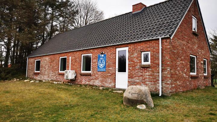 Viser spejderhytten i Tim. Rød murstenshus med hvide vinduer og dør. Sort tag og med spejderlogoet på muren.